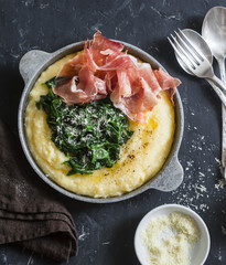 Polenta with butter spinach and prosciutto in a frying pan. Delicious lunch in a mediterranean style. On a dark background, top view.