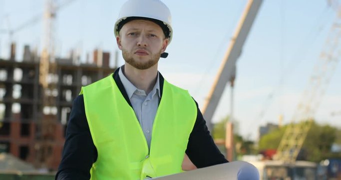 Young caucasian builder in white helmet on unfinished construction background holding the drawing and looking on the camera. Outdoor.