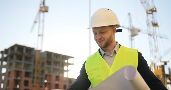 Handsome caucasian architect in white helmet standing on building background. Caucasian builder looking drawing and smiling. Outdoor.