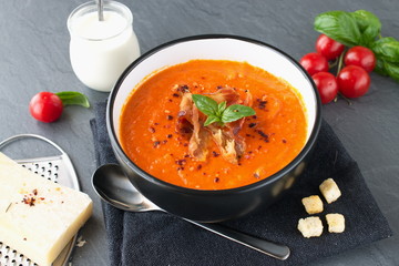 Thick tomato soup with basil and fried bacon in a black ceramic bowl on a grey abstract background. Healthy eating concept Fresh tomato soup with creme fraiche