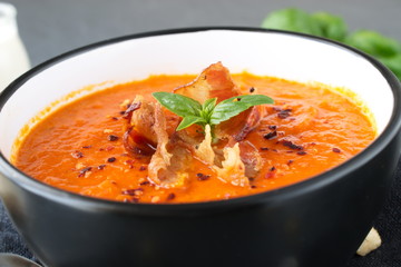 Thick tomato soup with basil and fried bacon in a black ceramic bowl on a grey abstract background. Healthy eating concept Fresh tomato soup with creme fraiche