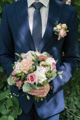 Beautiful lush wedding bouquet of white and pink peony and roses in groom's hands. Groom in suit with wedding bouquet outdoors