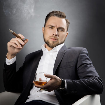 Man with cognac glass and cigar. Hair style, beard, suir and white shirt