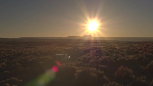 AERIAL LOW ANGLE: Black SUV car driving through vast bushy desert at stunning golden sunrise. All terrain vehicle on journey through desert landscape in sunny morning. Jeep at perfect sunset in desert