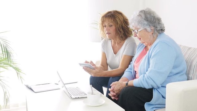 mature woman helping assisted elderly senior female with administrative procedures and paperwork on internet with a laptop computer at home