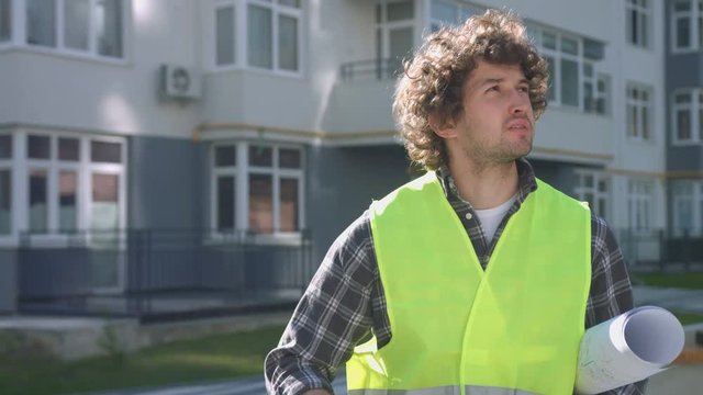 Young caucasian builder with black curly hair putting on head the yellow protective helmet, looking around on the background built house.