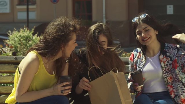 Pretty caucasian girl bragging about shopping to her friends. Three young women sitting on the bench outdoors. Attractive female teenagers looking inside the bag