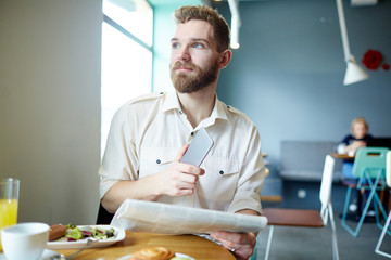 Serious broker with smartphone and newspaper eating in cafe