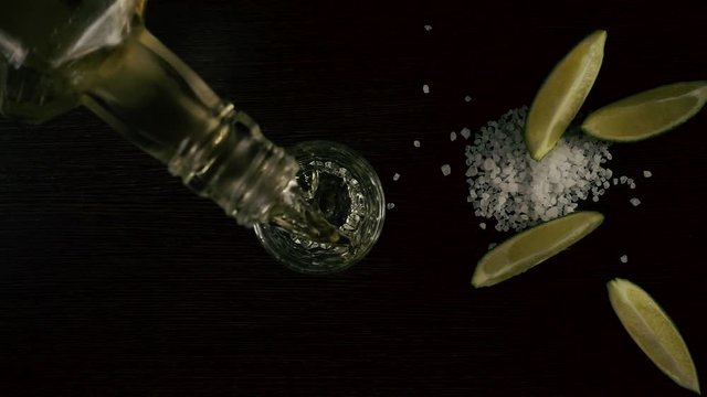 Slow motion. The bartender from the bottle will pour the tequila in a glass on the table and next to the lime and salt. Top view