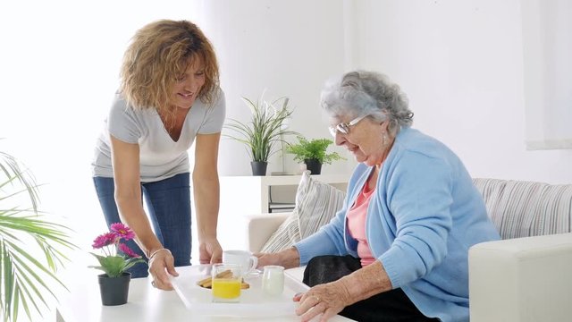 cheerful mature woman serving breakfast and taking care of elderly senior woman at home