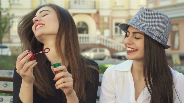 Cute young woman blowing soap bubbles on the bench. Two female friends having fun outdoors in the city. Close up of two pretty girls sitting against background of some public building