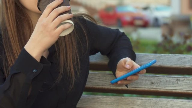 Attractive young woman drinking coffee on the bench outdoors. Close up of female hands holding smartphone and plastic cup. Brown haired girl taking a sip of hot drink