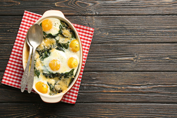 Baking dish with delicious cooked eggs Florentine and napkin on wooden background