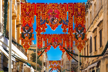 Naklejka premium Festively decorated street with banners for St Augustine Feast in the old town of Valletta, Malta. Flaming, arrow pierced heart - symbol of St Augustine