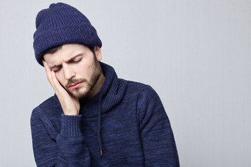 Stressed Caucasian hipster man having upset and frustrated look, suffering from bad headache, grimacing, squeezing his head with painful expression. Young male in blue hat feeling stress.