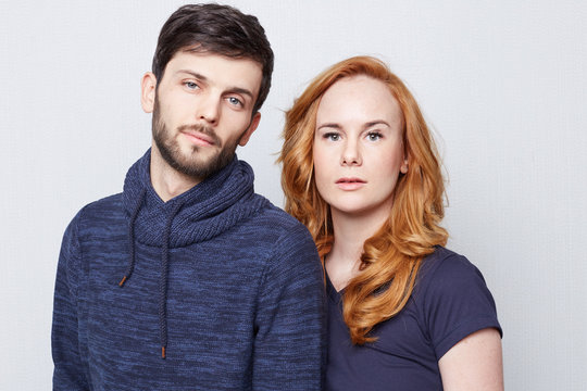 Portrait of two young beautiful Caucasian people wearing blue clothes posing on white background. Beautiful girl with long red hair looking confident while standing behind her handsome bearded man.