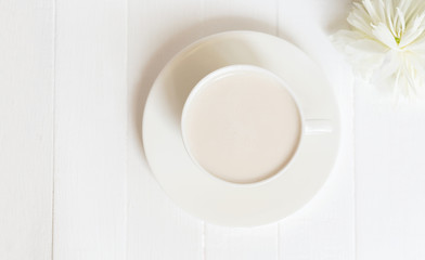 On a white wooden background there is a cup of coffee. Next to it is a white peony flower