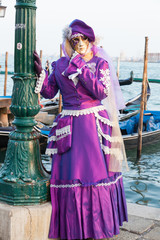 Obraz premium Venice 2017 Carnival, Italy. Woman in colorful purple dress and mask posing in front of gondolas