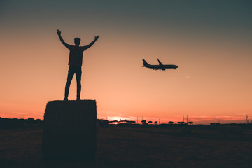 silhouette di un Ragazzo con le mani al cielo su una balla di fieno saluta il passaggio di un'aereo civile