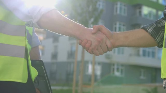 Portrait of hands of two caucasian builders. Builder holding tablet and shaking hand the builder on built house background.