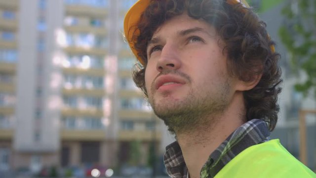 Portrait of side view of caucasian young builder with black curly hair in the protective helmet, looking up. Outdoor.
