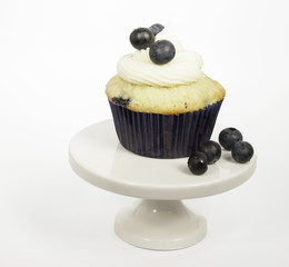 Cream cheese frosted blueberry muffin in dark blue paper with loose blueberries on a white pedestal against a white background.