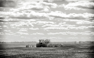 Obraz premium A tractor pulling a seeder and fertilizer tanks planting a crop in a black and white countryside summer landscape