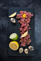 Tuna tartar with quail egg yolk, lemon and lime on a stone slate tray. Flat-lay on a dark metal background