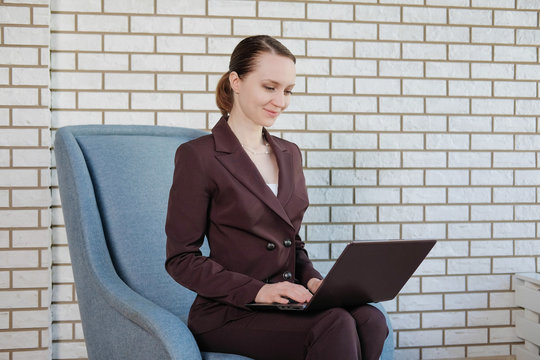 Smiling Businesswoman Working On Laptop In A Bright Office On The Background Of A White Brick Wall. Looking Away From The Camera. Girl In Elegant Office Suit.