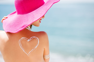 Back view on a beautiful woman in pink hat with sunscreen heart shape on her shoulder having a...