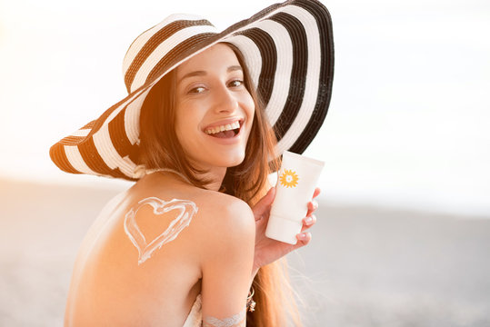 Beautiful woman in striped hat with sunscreen heart shape on her shoulder holding a sun cream bottle on the beach