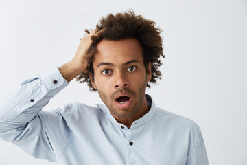 Obraz premium Confused hipster guy with African haitrstyle wearing formal clothes looking at camera with bugged eyes and dropped jaw touching his head with hand realizing his mistakes having shock and surprise
