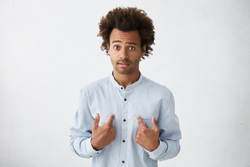 Startled handsome African businessman in elegant white shirt pointing at himself with hands trying to defence himself raising his eyebrows with great surprise. Man asking something about himself