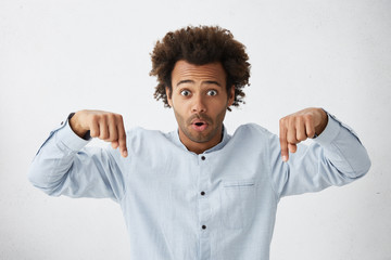 Cropped shot of shocked man with curly bushy hair and dark eyes wearing formal shirt pointing down with fingers looking with opened mouth at camera. People, emotions, feelings, body language concept © WHstudio Leushin N