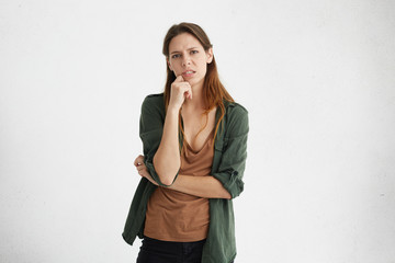 Upset woman with beautiful appearance wearing casual brown T-shirt and green jacket holding her hand on chin having tired and unhappy look. People, emotions, body language and lifestyle concept