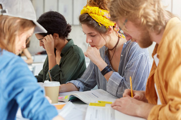 Youth, education and friendship concept. Group of clever creativ students sitting together in canteen surrounded with books and notebooks drinking coffee having concentrated and serious look