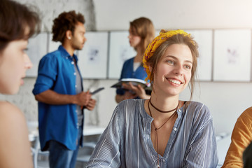 Obraz premium Smiling business lady wearing trendy shirt and yellow head bandage having pleasant smile while having business meeting with her partners isolated over working space. Communication, work concept