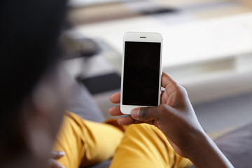 Indoor shot of hand of dark-skinned man holding cell phone with blank copy space screen for your text or advertising content while lying at couch at home searching internet. People and technology