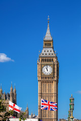 Big Ben with flag of England and United Kingdom against blue sky.