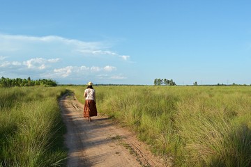 Naklejka na ściany i meble The girl in a brown skirt wearing a hat was walking on a dirt road. The blue sky is the backdrop