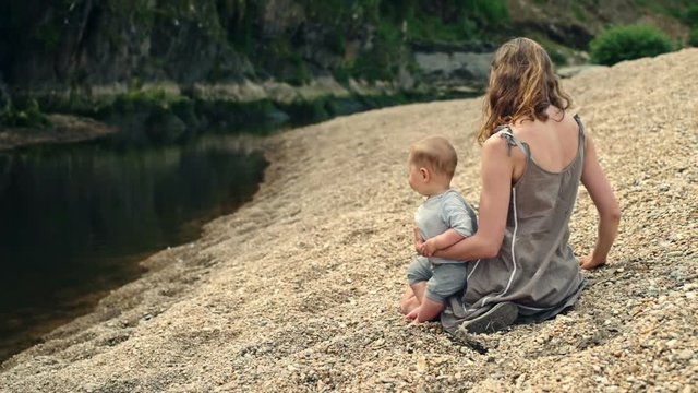 Young mother with baby sitting by river and throwing rocks