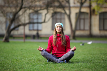 Smiling young woman meditating in the lotus sitting on the grass in the park, a healthy lifestyle, looking at the camera