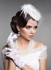 Fashion Retro elegant woman portrait. Wedding hairstyle. Brunette bride model present white hat, gloves and pearls jewelry accessories over gray studio background.