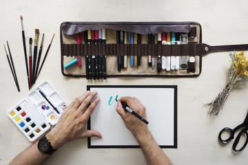 Top view on calligrapher hands writing with blue marker on blank paper on wooden table background with markers in leather case, dried yellow flowers, black scissors, brushes and watercolor palette.