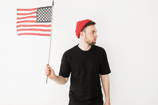 Young man in red hat and black t-shirt holds american flag and looks away, isolated on white background