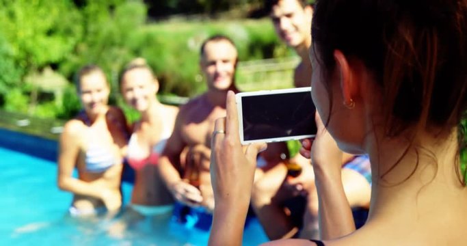 Smiling woman clicking photos of friends from mobile phone near poolside
