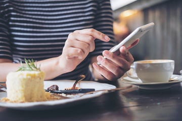 A woman using smart phone with cake and coffee cup on wooden table in restaurant