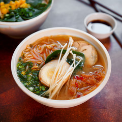Ceramic bowl of traditional asian ramen soup with noodles, spring onion, chicken, sliced egg and mushrooms served with wooden chopsticks on white table. Selective focus.