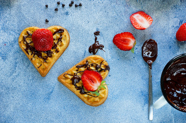 Belgian waffles in the shape of hearts with fresh strawberries and chocolate topping on blue table. Homemade healthy breakfast, top view