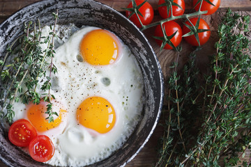 Fried eggs in a frying pan with herbs, cherry tomatoes, bread, olive oil and spices on rustic wooden background. Healthy breakfast, top view